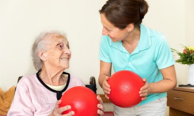 Physiotherapeutin und Patientin halten in einer Wohnung jeweils einen roten Ball